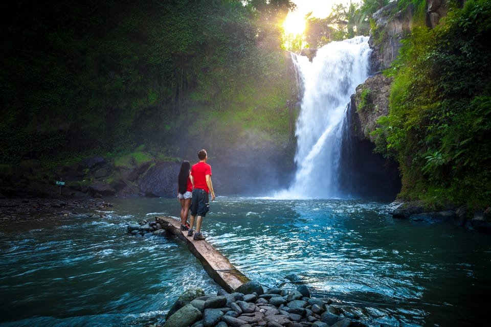 Uma pessoa de chapéu está em uma costa rochosa, de frente para a Cachoeira Tegenungan em Bali cercada por folhagens verdes e névoa. 