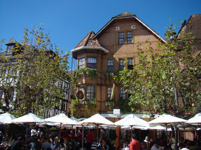 Prédios em estilo europeu numa rua do Capivari em Campos do Jordão, com mesinhas de madeira e guarda-sóis com pessoas sentadas