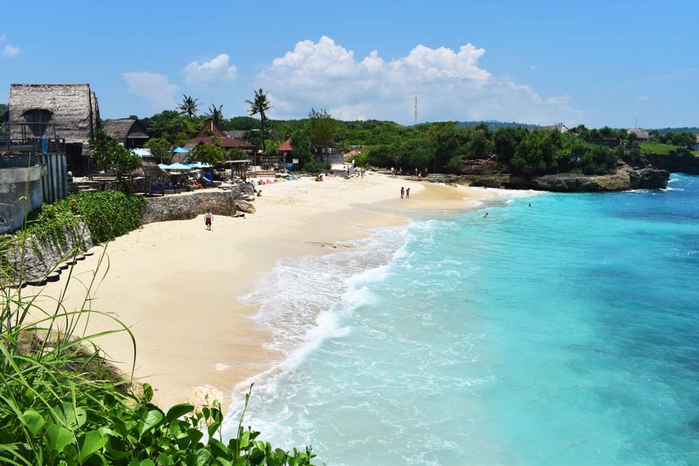 Foto tirada do topo de algum morro ou colina da Dream Beach em Nusa Lembongan. O mar, à direita, é azul e cristalino, a areia, no lado oposto, é clara. O céu está azul e com poucas nuvens.