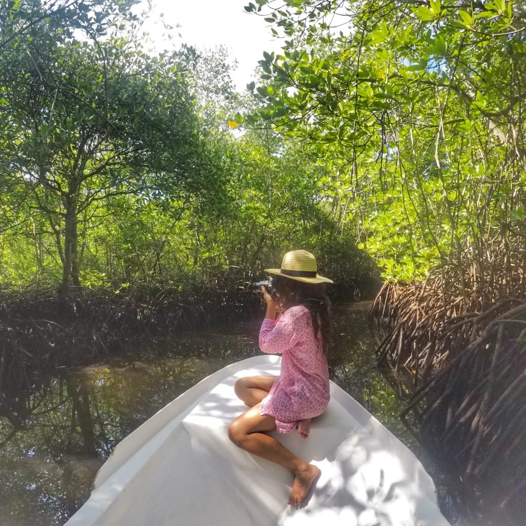 Mulher em um barco branco pequeno em um ago na Mangrove Forest, em Nusa Lembongan. Ela veste um vestido rosa e usa um chapéu, e está tirando foto dos manguezais à sua frente.