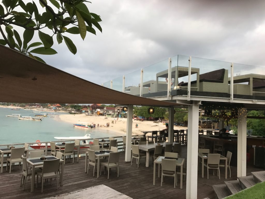 Foto do restaurante Ware Ware Rooftop Bar, mostrando um deck de madeira com várias mesas e cadeiras com vista para uma praia. Há um segundo andar no restaurante, com mais mesas ao ar livre.