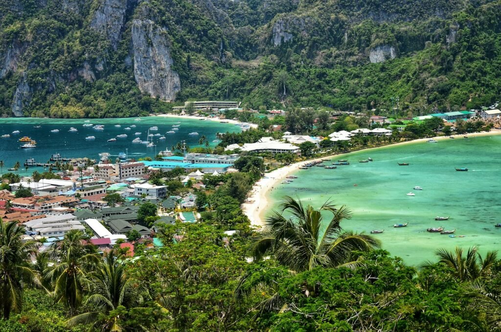 A imagem mostra uma vista aérea das Ilhas Phi Phi, localizadas na província de Krabi, na Tailândia. A paisagem é uma mistura de natureza exuberante e infraestrutura turística. No centro da imagem, há uma pequena vila costeira com edifícios de diferentes tamanhos e cores. A água do mar é clara e exibe diferentes tons de azul e verde, refletindo a luz do sol. Em ambos os lados há diversas embarcações, O céu está parcialmente nublado, com nuvens brancas esparsas, permitindo a passagem de luz solar que ilumina toda a cena com um brilho suave. Representa eSIM Tailândia.