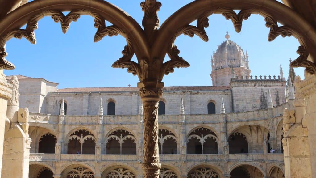 Arcos de pedra emolduram a vista do Mosteiro dos Jerônimos, um claustro histórico ornamentado com entalhes detalhados e uma alta torre abobadada sob um céu claro, que pode ser observado com o eSIM Lisboa.
