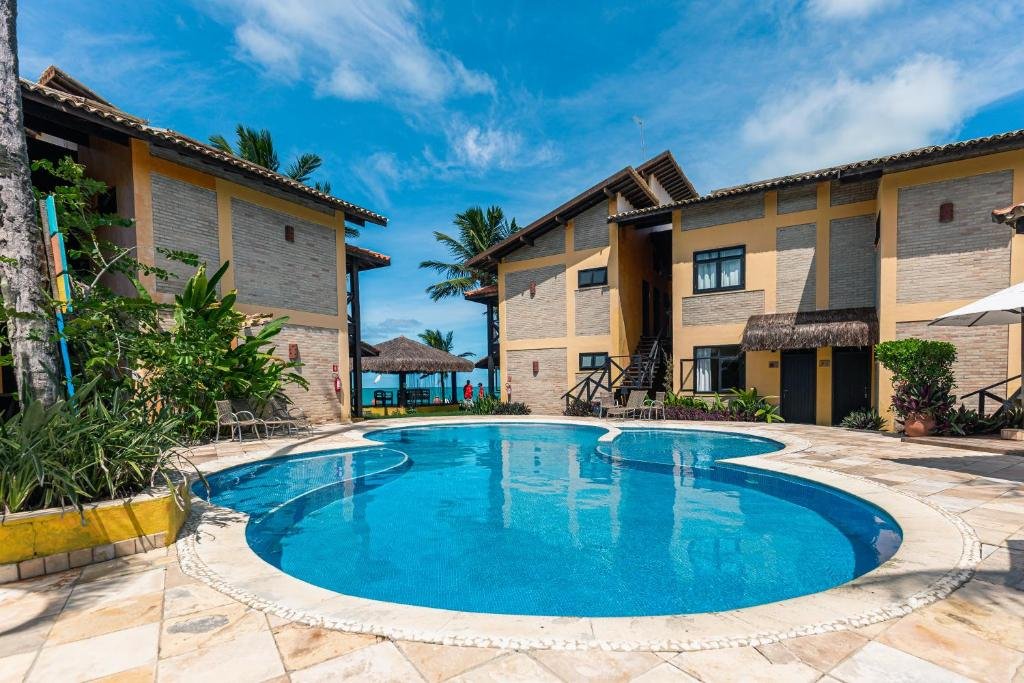 Piscina ao ar livre do Anauí Pousada. Do lado esquerdo e do lado direito a pousada, no fundo um guarda-sol de palha e a praia.