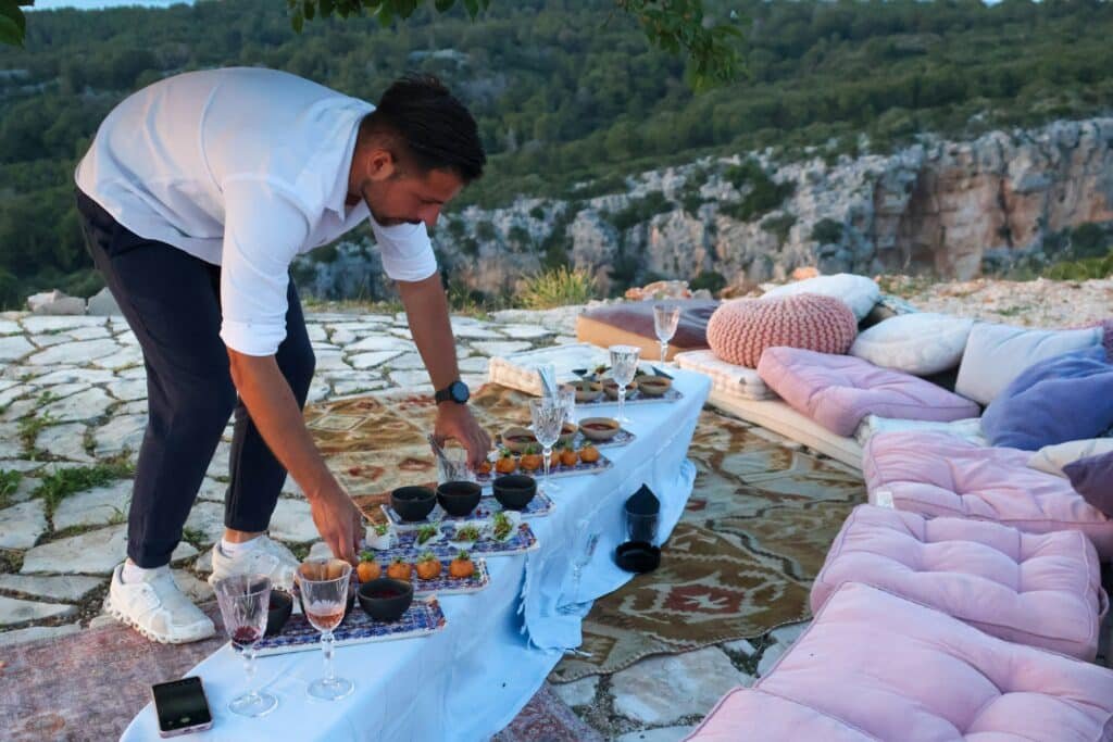 Um homem arruma comida e bebidas em uma mesa baixa colocada em um pátio de pedra ao ar livre em Hvar com vista panorâmica. Assentos confortáveis cercam a mesa.