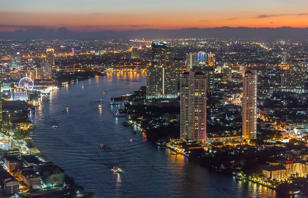 Vista aérea de Bangkok ao anoitecer com um rio, arranha-céus iluminados e luzes da cidade ao redor sob um céu colorido. Imagem para ilustrar post sobre o que fazer na Tailândia.