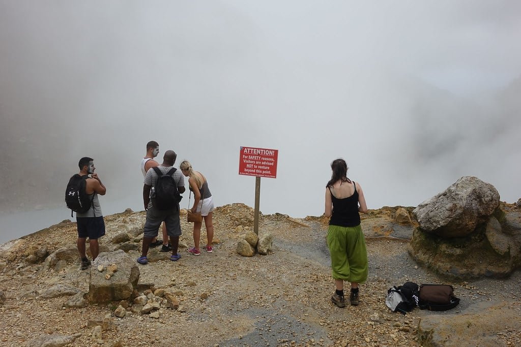 Cinco pessoas estão perto de uma placa de alerta vermelha em uma área rochosa e úmida, com baixa visibilidade, e uma mochila no chão no Boiling Lake. Imagem para ilustrar post sobre eSIM Dominica.