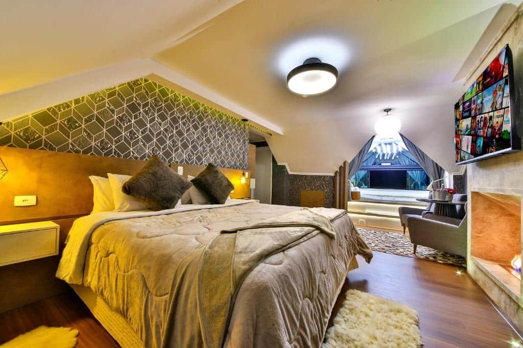 Quarto do Caminho das Pedras Pousada de Charme com cama de casal, tapetes, chão de madeira, lareira, banheira de hidromassagem com uma ampla janela