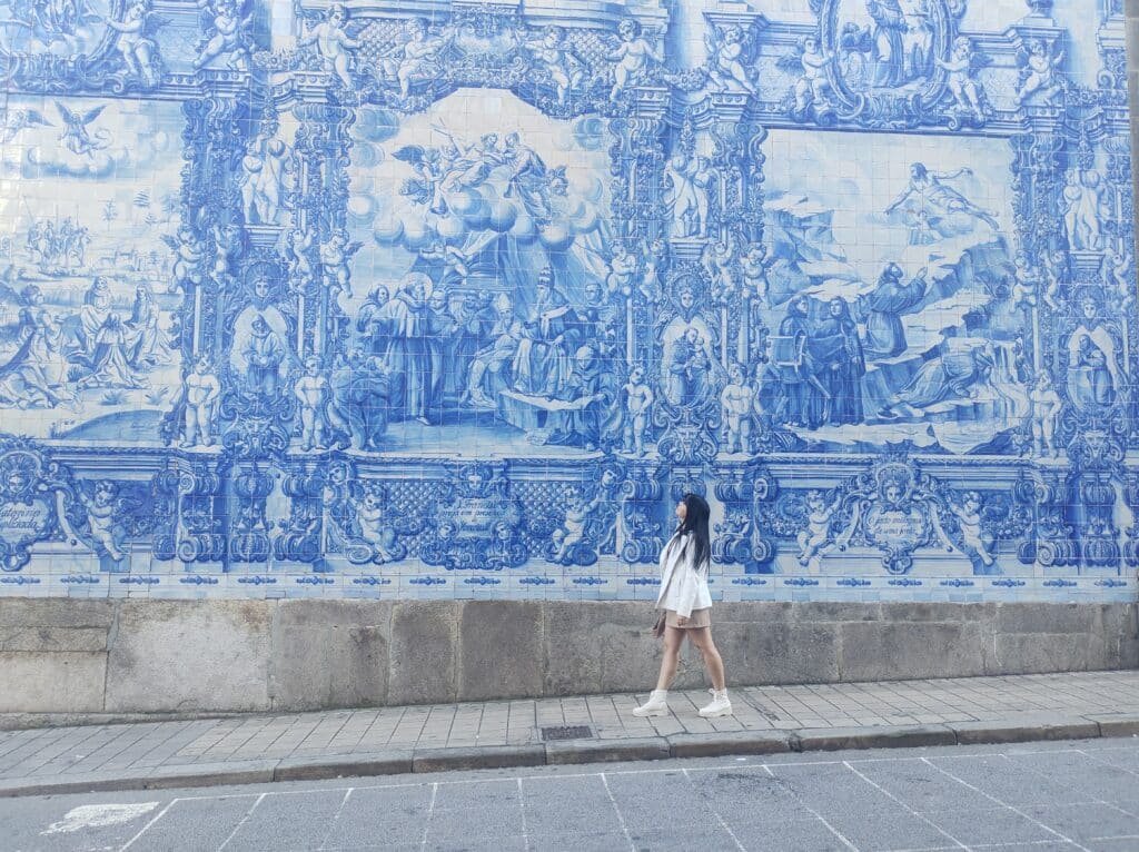 Uma mulher caminhando pela rua com o mural de azulejos ao fundo.