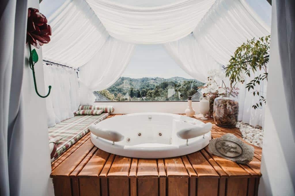 Banheira do Chateau La Villette cercado por janelas de vidros e panos brancos, o deck é de madeira com vasos de plantas e almofadas
