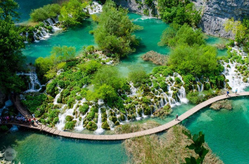 Foto aérea do Parque Nacional dos Lagos de Plitvice, mostrando o enorme lago que é cortado por árvores cachoeiras, montanhas  uma ponte de madeira.