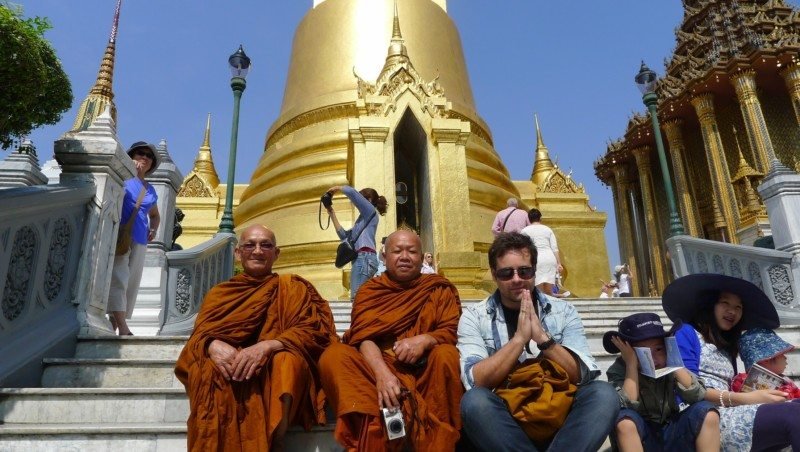 Dois monges e o nosso colunista sentados nos degraus do Grand Palace em frente a um pagode dourado. Uma mulher ajusta uma câmera ali perto. Dia claro e ensolarado.