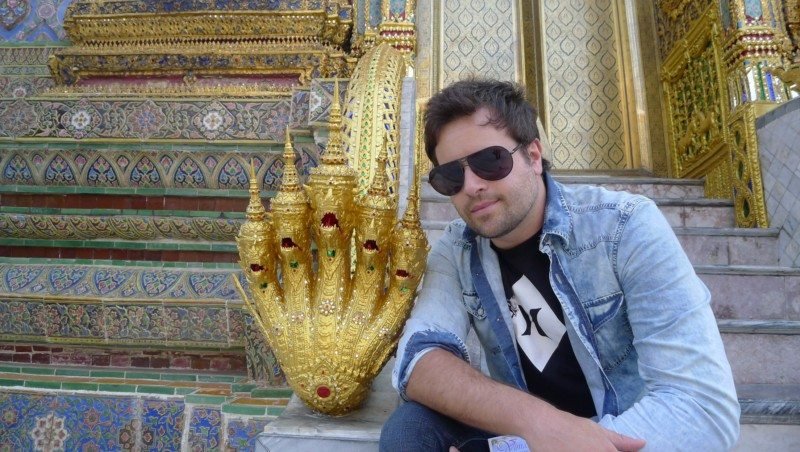 Na Tailândia, uma pessoa usando óculos escuros e uma jaqueta jeans está sentada nos degraus do Grand Palace perto de uma estrutura dourada ornamentada com desenhos complexos.