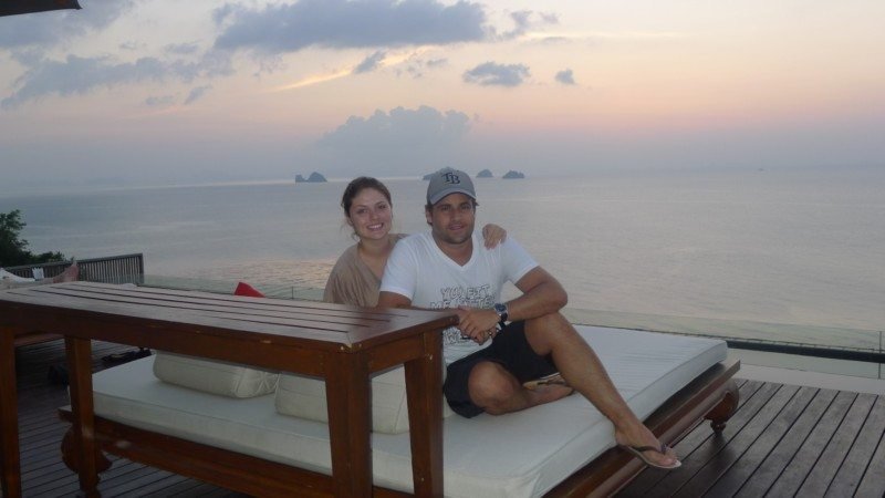 Um casal está sentado em um deck com almofadas, com vista para o oceano ao pôr do sol na Tailândia. O céu está nublado e a água está calma.