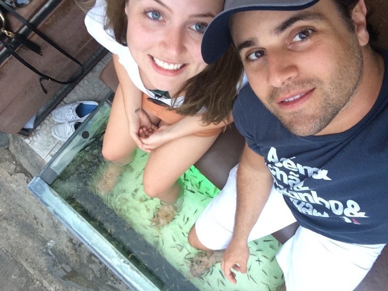 Uma mulher e um homem estão fazendo uma pedicure com peixes, com os pés submersos em um tanque de vidro cheio de pequenos peixes.