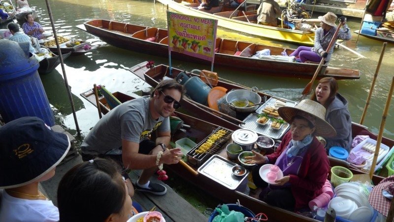 Nosso colunista ao lado de uma mulher vendendo comida de um barco em um movimentado mercado flutuante. Outros vendedores e barcos são visíveis ao fundo.