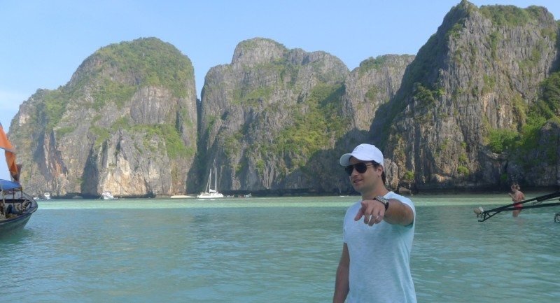 Na Tailândia, um homem com boné branco em pé em águas rasas com grandes penhascos rochosos e barcos ao fundo, na Ilha de Kho Phi Phi.