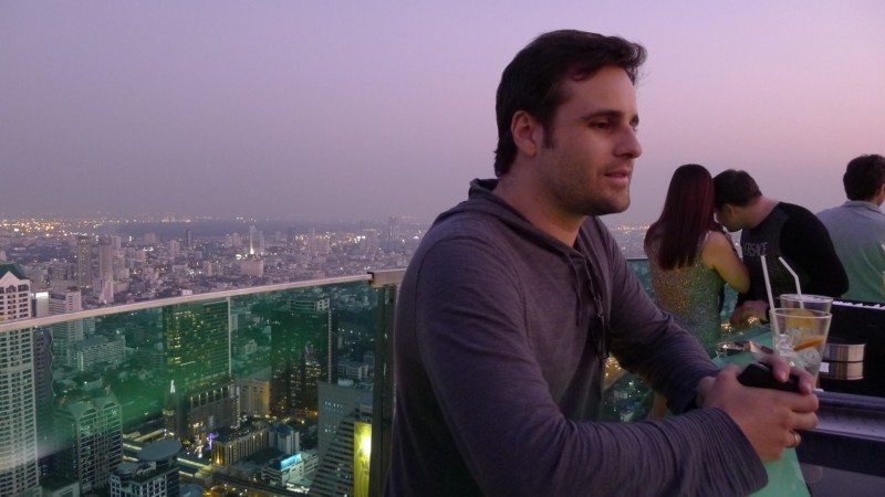 Na Tailândia, um homem está na cobertura do Sky Bar com vista para uma paisagem urbana ao anoitecer, segurando uma bebida, na Tailândia. Há outras pessoas ao fundo.