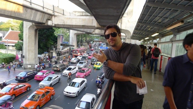 Uma pessoa se apoia em um corrimão em Bangkok, com vista para uma rua movimentada com carros e ônibus, abaixo de uma estrutura de concreto elevada.