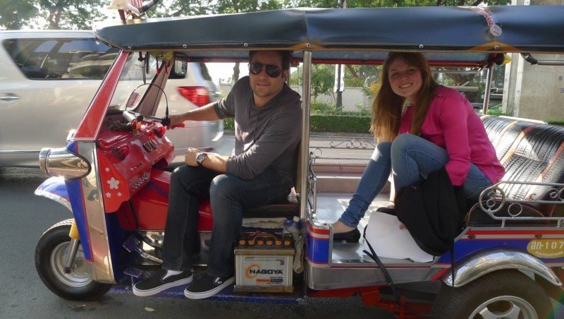 Duas pessoas estão sentadas em um tuk-tuk colorido em uma rua. O homem está no assento do motorista, e a mulher está sentada atrás.