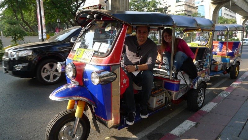Duas pessoas sentadas em um tuk-tuk estacionado em uma rua da cidade de Bangkok, na Tailândia, com árvores e prédios ao fundo.