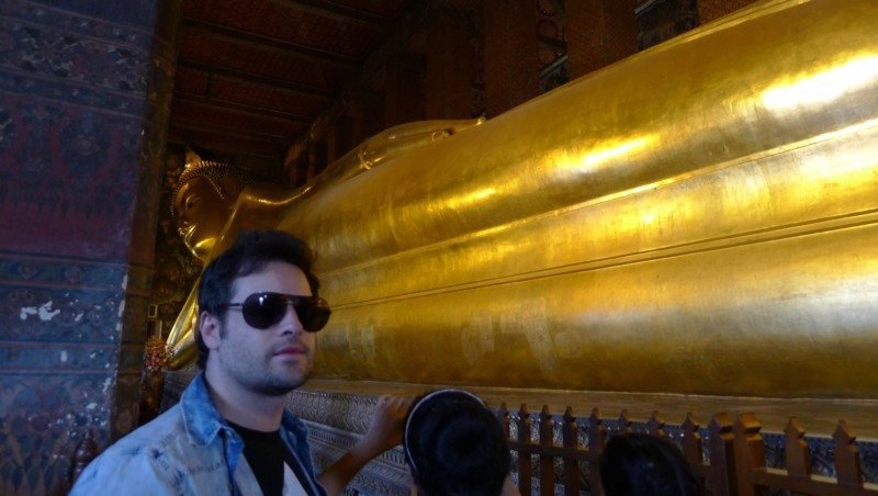 Nosso colunista usando óculos escuros está em frente a uma grande estátua dourada de Buda reclinado dentro de um templo mal iluminado, no Wat Pho. Imagem para ilustrar post sobre o que fazer na Tailândia.