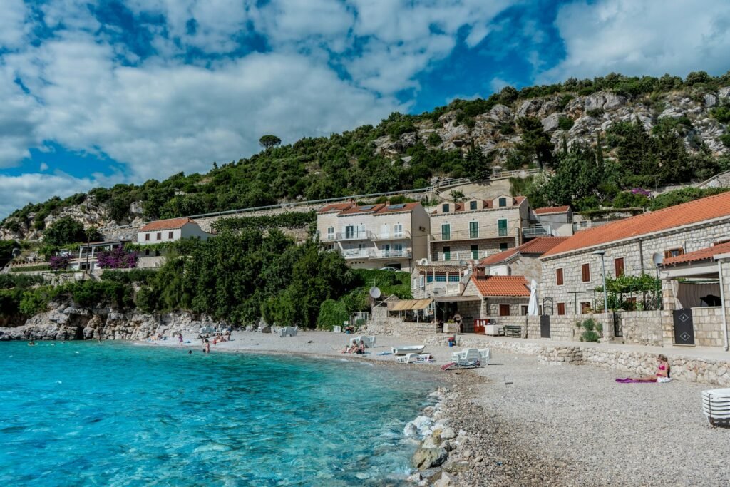 Uma praia de seixos com águas azul-turquesa em Dubrovnik, com pessoas tomando sol e construções de pedra na encosta de uma colina sob um céu parcialmente nublado. A foto faz parte do post de eSIM Croácia.