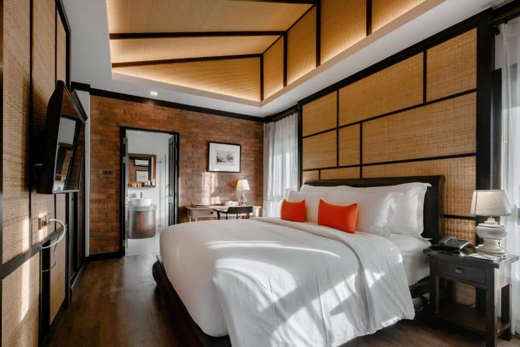 Um quarto aconchegante com uma cama grande, roupa de cama branca, travesseiros laranja, piso de madeira e uma parede de tijolos com TV.