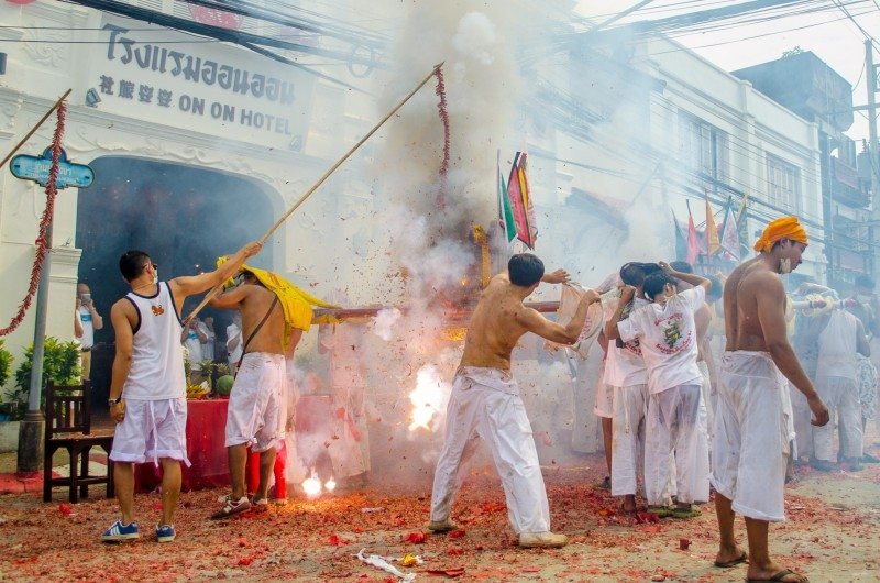 Um grupo participa de um festival  em Phuket com fumaça e fogos de artifício do lado de fora de um prédio. Os participantes vestem roupas brancas e carregam bandeiras. Imagem para ilustrar post sobre o que fazer na Tailândia.