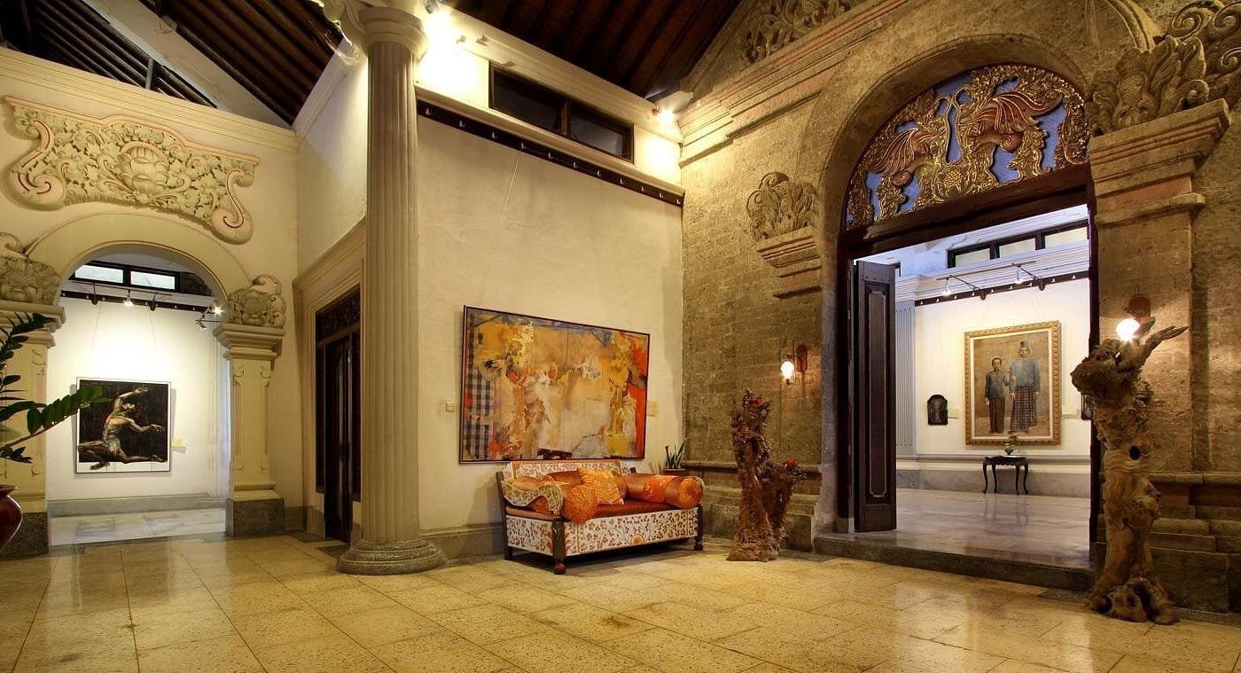 Uma sala espaçosa e repleta de arte, com um arco de pedra, pinturas nas paredes, um sofá, pilares ornamentados e uma escultura à direita. É a Galeria de Arte de Agung Rai (ARMA) em Ubud.