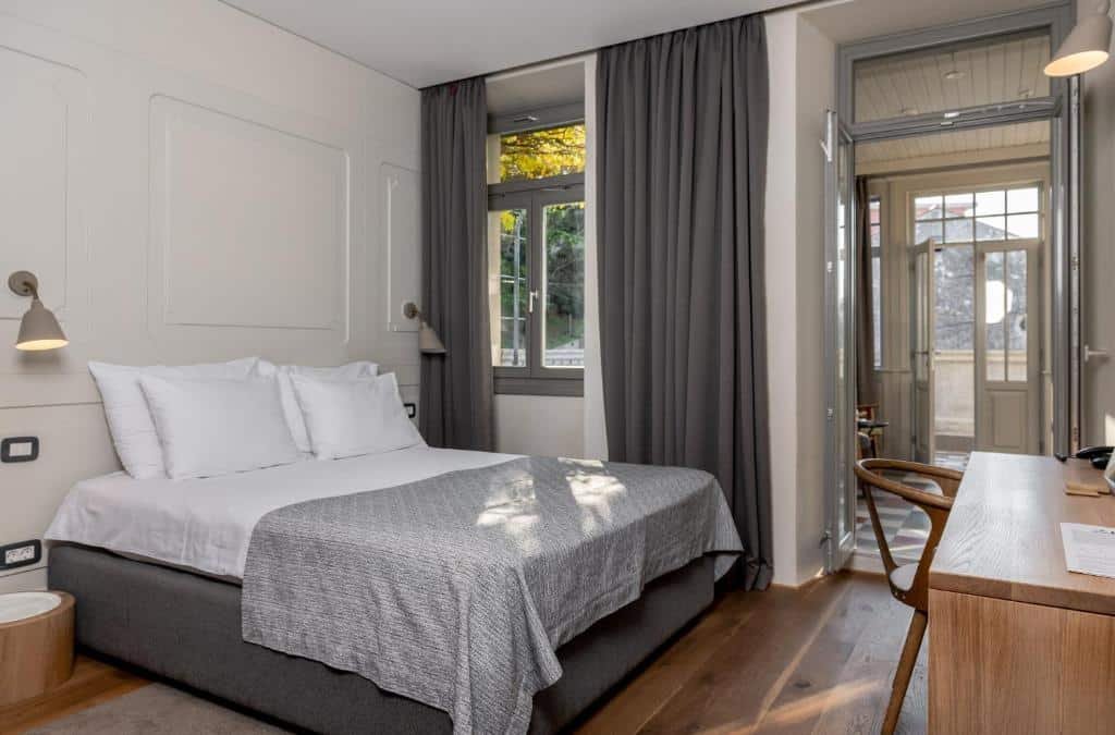 Quarto do Hotel Peteani bem iluminado com cama de casal, cortinas cinza, piso de madeira, escrivaninha e uma porta que dá acesso a uma pequena varanda com vista.