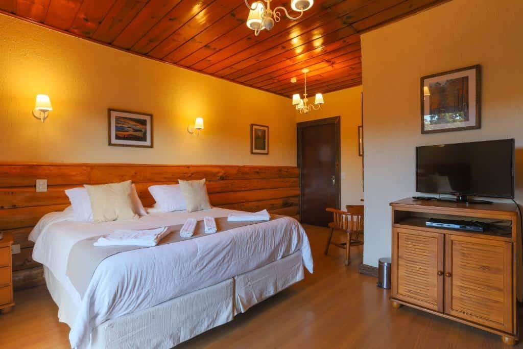 Quarto no Hotel Savoy Excellence com chão de madeira, cama de casal, uma cabeceira de madeira, com lustres, um móvel de madeira com uma televisão em cima, para representar onde ficar em Campos do Jordão