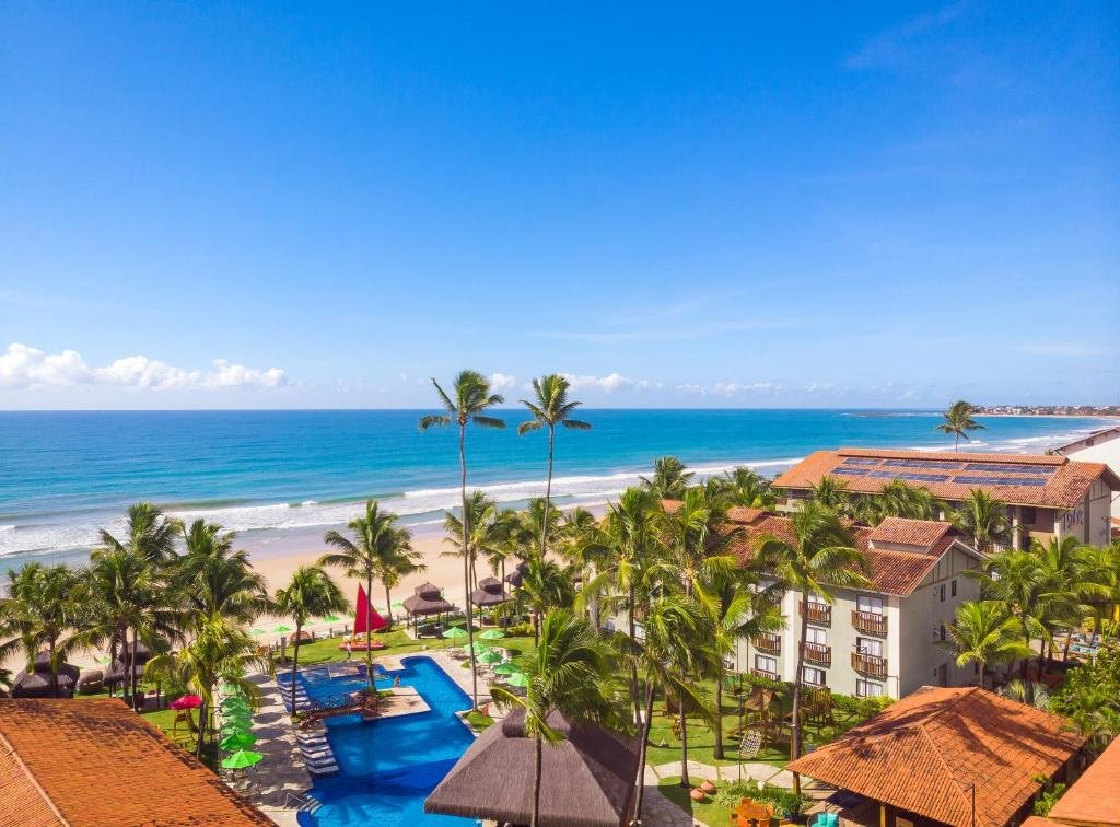Vista de cima do Hotel Solar Porto de Galinhas. O prédio nas laterais, no meio coqueiros e uma piscina, no fundo a areia e o mar.