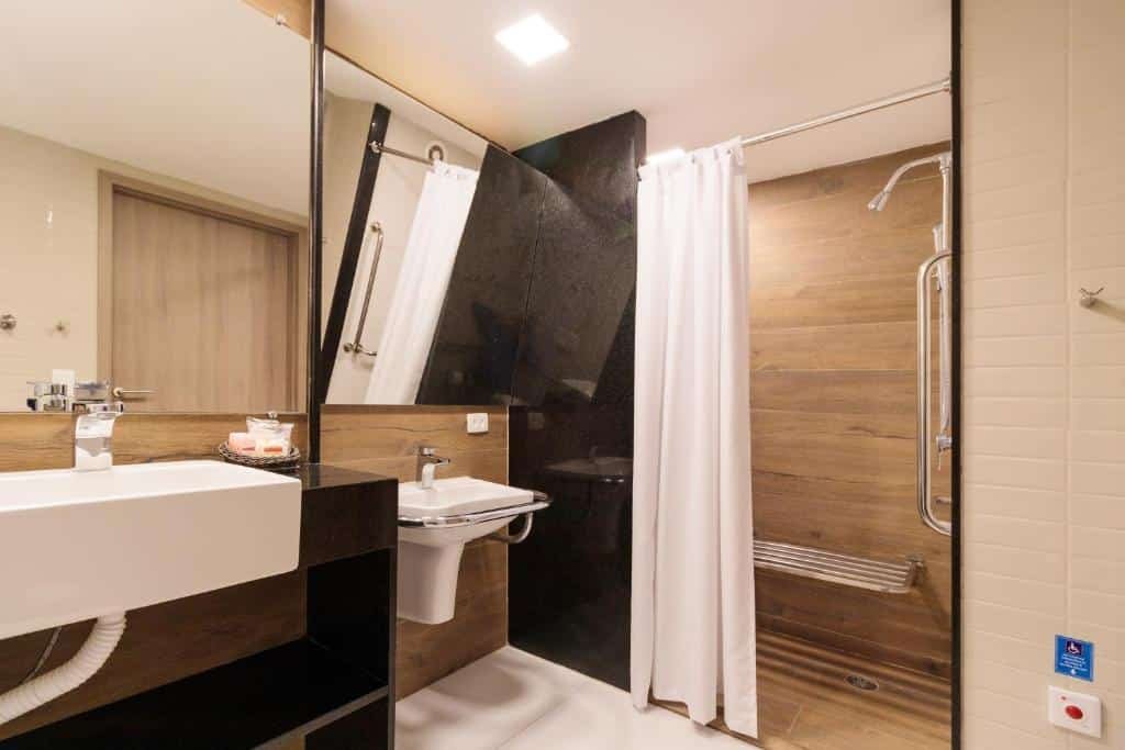 Banheiro com acessibilidade do Hotel Solar Porto de Galinhas. Do lado direito no fundo um box com cadeira e barras de apoio, no meio uma pia rebaixada com barras e espelho inclinado para baixo. No lado esquerdo uma pia alta com espelho.