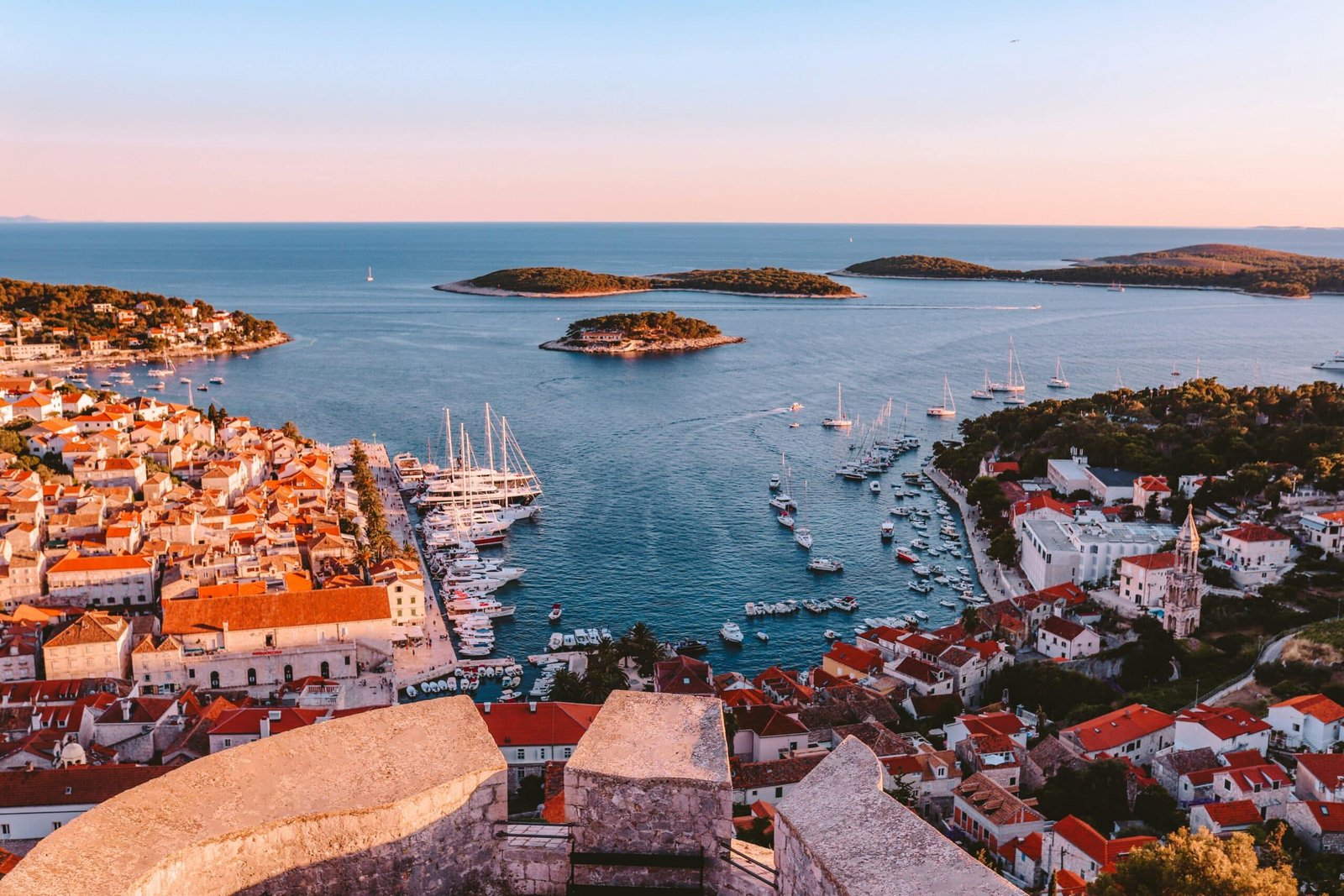 Vista aérea da cidade costeira de Hvar com prédios de telhados vermelhos, marinas e várias pequenas ilhas em um mar azul calmo ao pôr do sol. Ilustra o post de Roteiro para Hvar.