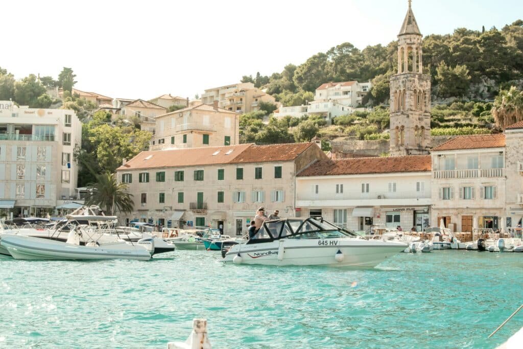 Barcos estão atracados em um porto ensolarado de Hvar Town com edifícios e uma igreja com uma torre alta ao fundo.