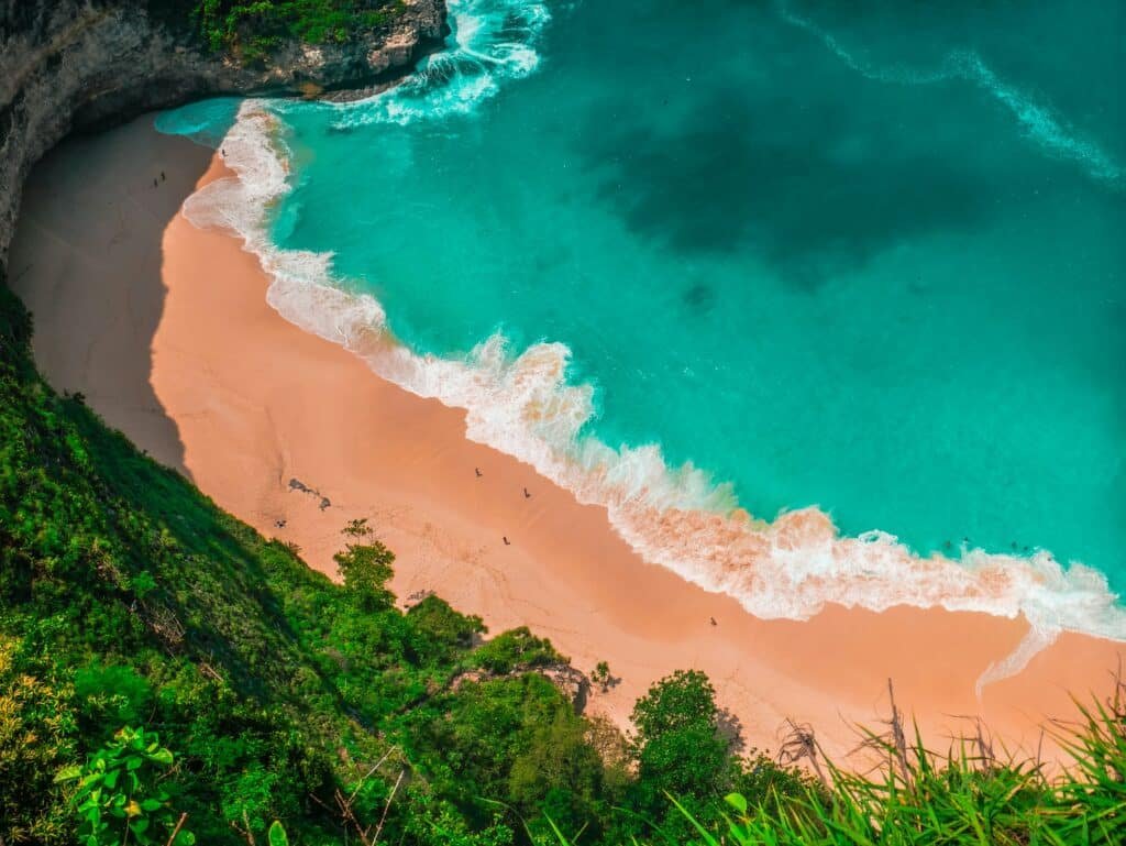 Vista aérea de Kingking Beach com águas azul-turquesa, ondas brancas e uma costa arenosa, cercada por penhascos e vegetação verde. Representa Bali.