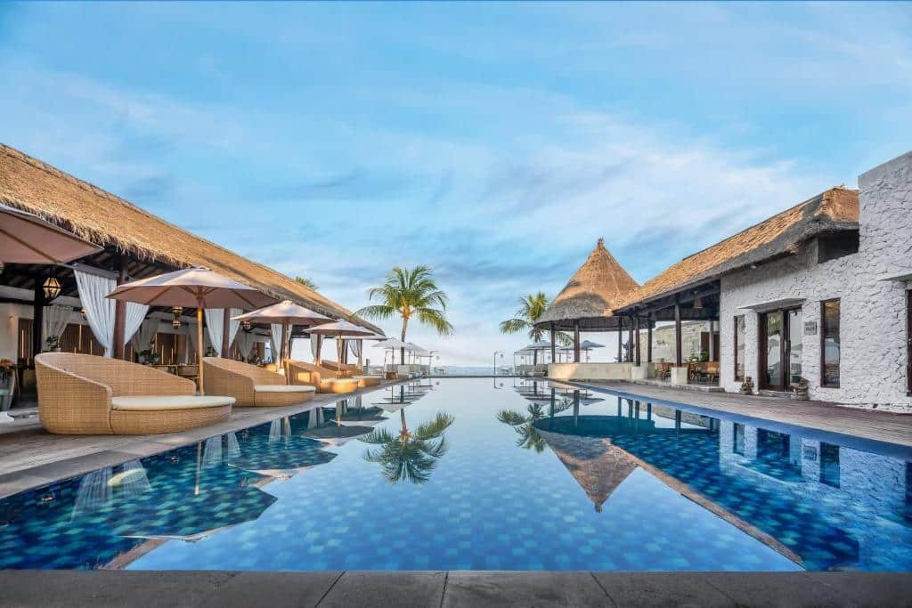 Uma luxuosa piscina do Lembongan Beach Club & Resort com espreguiçadeiras de vime, palmeiras e edifícios com telhados de palha sob um céu azul claro.
