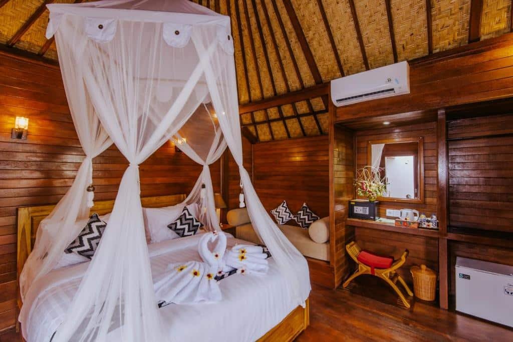 Um aconchegante quarto do Lembongan Small Heaven Bungalow de madeira dispõe de uma cama de dossel com toalhas de cisne e pétalas de flores, uma penteadeira com espelho e uma pequena área de estar.