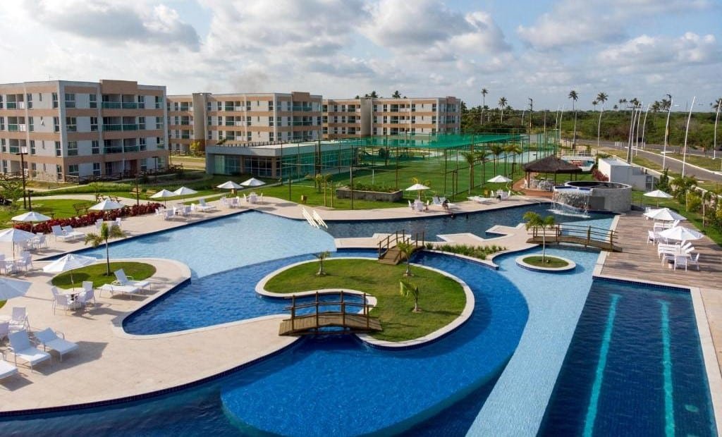 Vista da área externa do  Muro Alto Condomínio Clube. Uma piscina grande no meio, com pontes, ilha de jardim, e ao redor cadeiras de tomar sol. No fundo um campo, e os apartamentos.