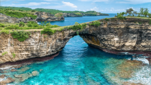 Um arco de rocha natural em Nusa se estende sobre águas cristalinas azul-turquesa, cercado por penhascos e arbustos verdes sob um céu parcialmente nublado. Representa Bali.