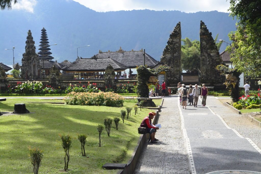 Entrada com um caminho pavimentado e jardim à esquerda até o templo Pura Ulun Danu Bratan, com vista para montanhas ao fundo.  Está ilustrando o post sobre o que fazer em Ubud.