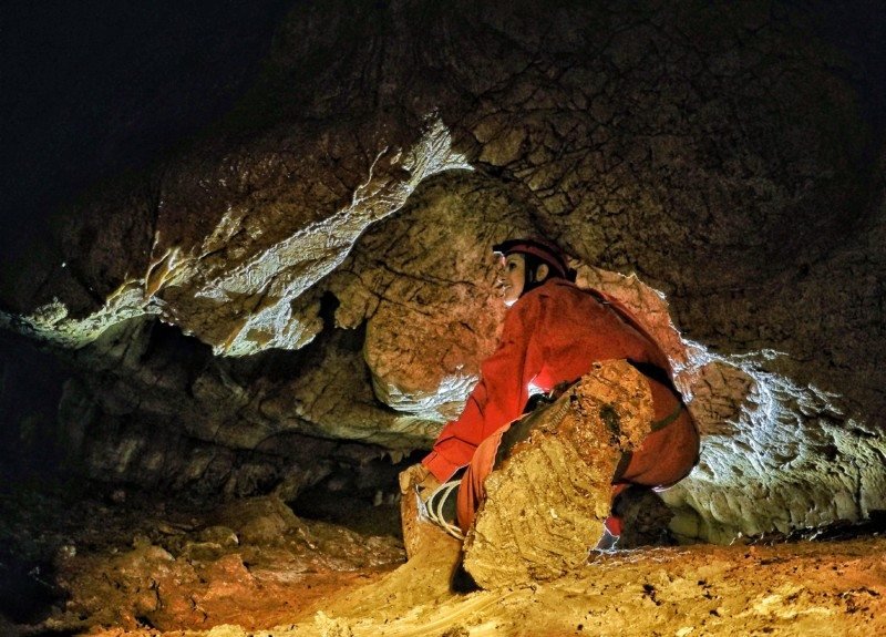 uma mulher agachada usando um macacão vermelho e equipamentos de escalado dentro da gruta de baredine