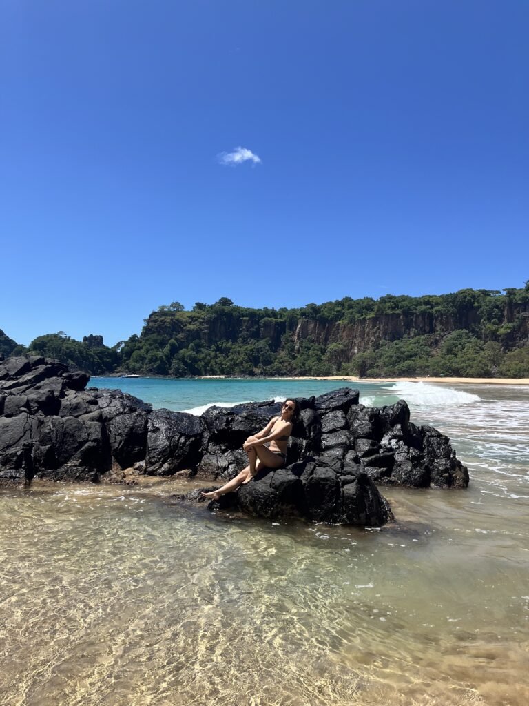 Uma pessoa sentada em pedras pretas na praia com águas cristalinas, uma praia de areia e penhascos verdes sob um céu azul.