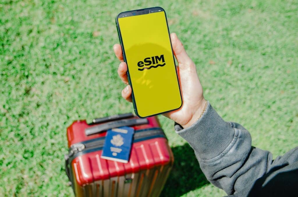 Uma pessoa segura um smartphone com a inscrição "eSIM" em uma tela amarela, acima de uma mala vermelha e um passaporte, na grama verde. Representa eSIM Tóquio.
