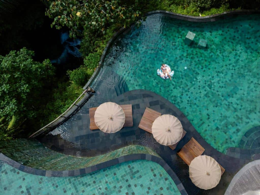 Vista aérea de uma piscina do Aksari Resort Ubud by Ini Vie Hospitality com mosaicos, cadeiras de piscina, guarda-sóis e uma pessoa flutuando em um inflável na água. Representa Bali.