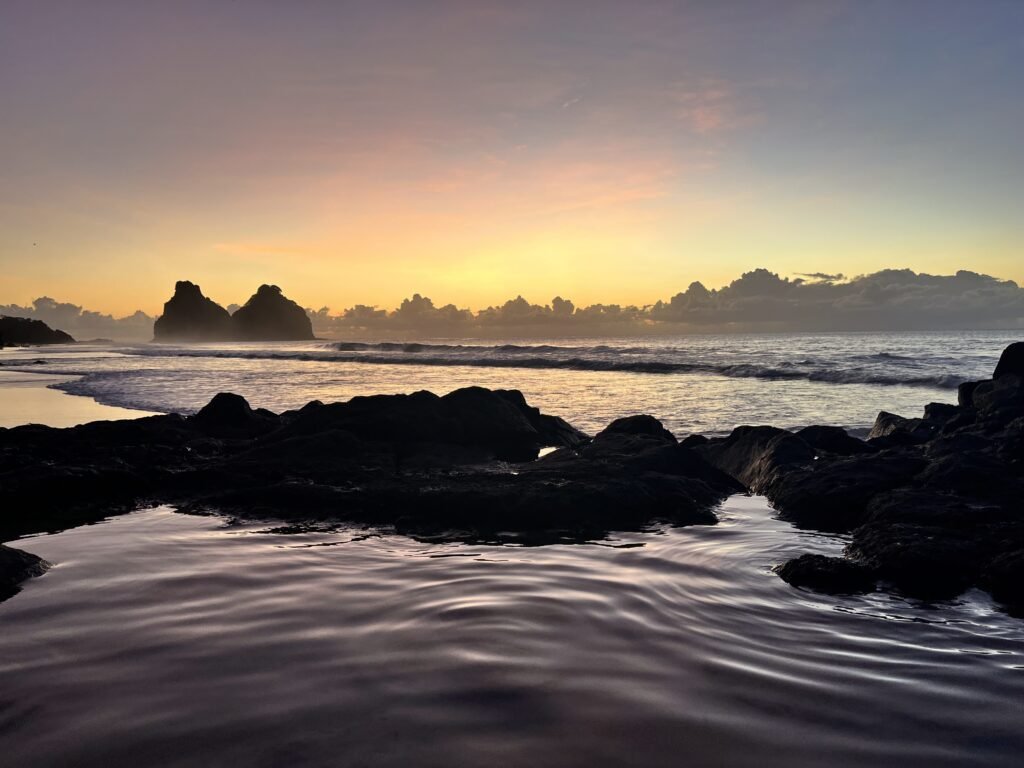 Litoral rochoso ao pôr do sol com águas calmas, silhueta de rochas e nuvens no horizonte sob um céu colorido.