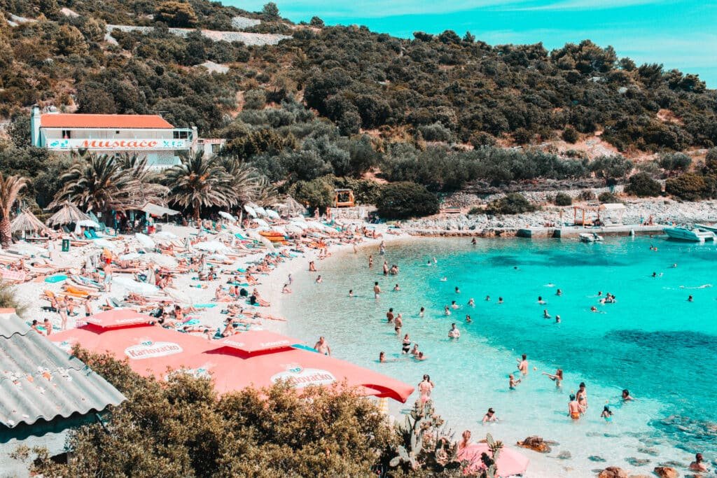 Foto da praia Pokonji Dol, mostrando várias pessoas dentro do mar e uma faixa de areia branca à esquerda com mais pessoas deitadas em suas toalhas e sentadas debaixo de guarda-sóis. No fundo da imagem há muitas vegetações.
