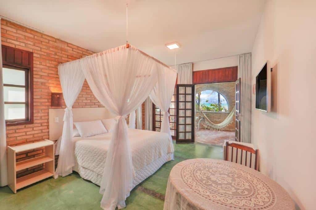 Quarto da Pousada Ecoporto. Uma mesa redonda e uma televisão do lado direito. Do lado esquerdo uma cômoda, e uma cama de casal. No fundo, a porta do quarto para a varanda com rede e vista para o mar. Foto para ilustrar post sobre pousadas em Ipojuca.