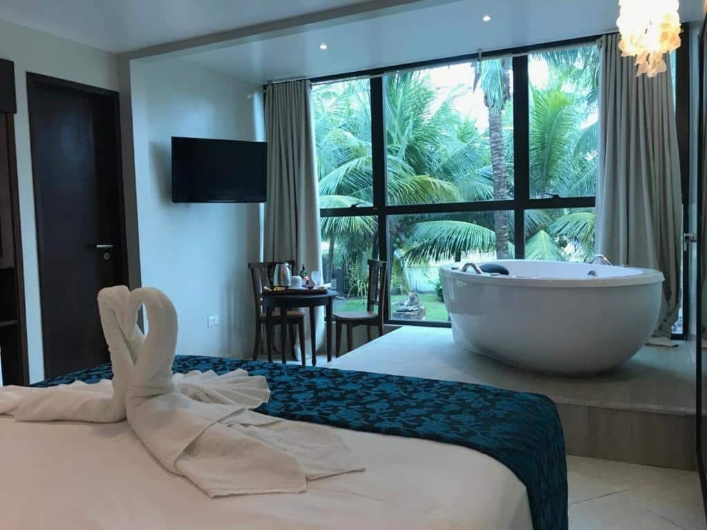 Quarto da Pousada Maracabana Spa. Uma piscina de casal no lado esquerdo, de frente uma televisão, uma mesa com duas cadeiras e a banheira de hidromassagem. Atrás uma parede de vidro com vista para o jardim. Foto para ilustrar post sobre pousadas em Ipojuca.